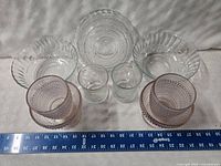 Top view of all glass items laid out on a white surface with measuring tape showing size, includes clear glass bowls and tumblers along with pink hobnail tumblers.