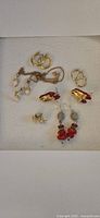 Photo showing all jewelry items including multiple pairs of gold-tone earrings, a gold-tone necklace with white leaf-shaped pendants, and matching red and white beaded earrings and necklace set.