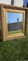 Full view of large decorative gold gilded frame mirror placed outside on grass showing reflective glass and ornate carved frame details.