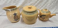 Front view of ceramic jug, lidded jar and teapot showing decorative floral and pineapple designs