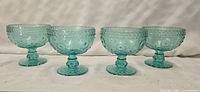 Front view of four teal glass footed bowls with embossed floral pattern arranged in a row.