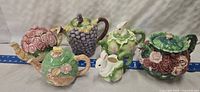 Six vintage ceramic teapots shown in various floral and fruit designs, one handle detached.