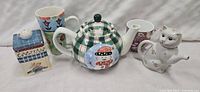 Five ceramic kitchen items including two teapots, two mugs, and one sugar packet holder arranged on a light surface.