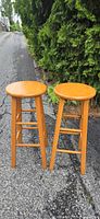 Photo of two wooden bar stools on pavement in front of green shrubbery, showing front and side view.
