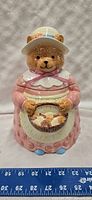 Front view of the ceramic teddy bear cookie jar wearing a pink dress and a white cap, holding a basket painted with cookies.