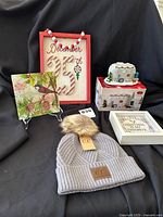 Lot items grouped on black cloth: grey beanie, decorative plate, framed sign, Christmas trailer figurine in box, and wooden framed signs
