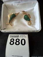 Two gold-tone costume brooches in white cushioned box; one shaped like a swordfish and the other a seahorse, both adorned with green stones.