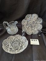 Set of two cut crystal plates and one pitcher displayed together on stands against a dark background showing star pattern and scalloped edges of plates and pitcher handle and spout