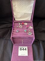 Front view of the purple velvet jewelry box opened showing the 5 rings and 2 pendants inside arranged in rows.