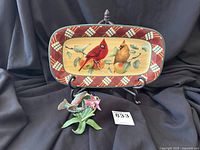 Colorful rectangular Lenox tray featuring a male and female cardinal on a branch with a red and white plaid border, displayed upright on a stand next to a green hummingbird statue with pink flower and foliage.