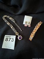 Photo showing all three items in the lot: the silver and gold-tone purple pendant necklace, pink earrings on a white earring holder, and amber stone bracelet.
