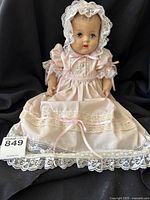 Full view of the porcelain baby doll wearing a long pink lace-trimmed dress and bonnet, seated against black background.
