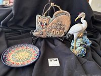 Photo of three pottery items against black cloth background: ceramic cats wall hanging, Polish pottery plate, and white stork figurine vase.