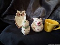 Image showing owl spoon rest, clown figurine, laughing pig figurine, and small yellow cup arranged on a black fabric background.