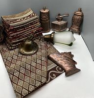 Photo showing the folded tapestry runner with matching napkins and several bronze-colored decorative items including a frosted glass light globe and metal candle holders or grinders.