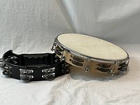 Two tambourines together: round, tuneable tambourine with drumhead and metal tuning lugs, and black plastic half moon tambourine with metal jingles.