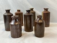 Group photo of 7 antique stoneware ink bottles in varying heights and shapes, dark brown glazed finish.