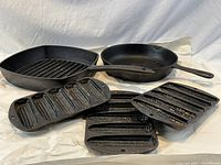 Five cast iron cookware items arranged on white paper including 3 corn trays, 1 square ridged grill pan, and 1 round skillet with handle