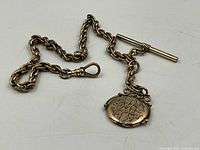 Full view of the gold filled twisted link fob chain with attached engraved oval locket and bar toggle clasp.