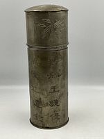 Front of pewter cylindrical brush box with etched bamboo on lid and Chinese characters on the body.