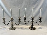 Pair of three-branch silver plated candelabras with white candles shown from front on white draped backdrop.