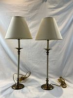 Pair of two tall stick table lamps with cream-colored shades and brass accent bases. Lamps have cords and are shown upright on white background.