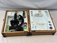 Open wooden case showing the microscope, colored lens filters, and prepared slide box inside foam insert.