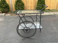 Full view of vintage wrought iron tea caddy bar cart showing black frame, glass top shelf with ornate border, two large spoked wheels, and handle.