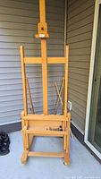 Front view of full wooden easel showing adjustable mast, lower shelf and wheels