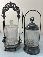 Two antique cut glass pickle castors side by side in decorative metal stands with lids and handles, small fork visible on one stand.