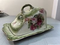 Front and side view of covered faience ironstone cheese dish with detailed floral pattern on lid, green shaded base with gold trim, and curved handle shaped like a leaf stem