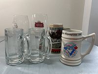 View showing seven beer glasses and steins including a white ceramic Toronto Blue Jays 1993 commemorative stein, a detailed Anheuser-Busch stein and clear glass Stella Artois glasses along with smaller glass mugs.
