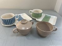 Wide shot showing two soup cups with attached square saucers, one with green leaf pattern and one with blue leaf pattern, alongside brown Melmac cream and sugar set.