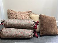 Five fabric cushions shown stacked, including two square pillow cushions and three patterned bolsters with ribbons.