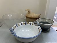 Image of four kitchenware items: a large ceramic bowl with blue floral leaf pattern and rope handles, a duck-shaped casserole dish, a frosted glass bowl, and a two-tone ceramic pot with saucer.