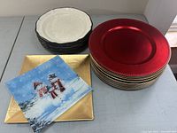 Six cream-colored Mikasa dinner plates stacked, next to red and gold charger plates and two decorative serving plates.