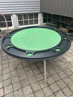 Front angle view of the round poker table showing green felt center and black plastic ring with cup holders and chip slots, foldable metal legs visible.