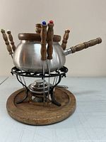 Side view of the fondue pot set showing the stainless steel pot on black metal stand with burner, wood base, and six wood-handled forks with colored tips inserted around the pot.