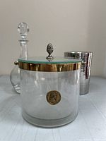 Clear glass ice bucket with brass rim and emblem, glass wine carafe with stopper, hammered metal wine chiller all displayed