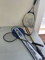 Four rackets on a light surface including two squash rackets, one tennis racket, and one badminton racket with Yonex cover.
