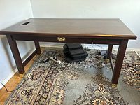 Wide angle of wood desk showing surface, legs, and drawer with gold handle. Desk placed on patterned rug.