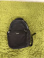 Front view of the black backpack placed on a green plush carpet, showing the front pocket and main compartment.