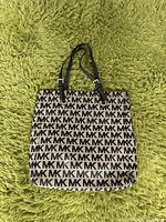 Michael Kors tote bag on green carpet background showing front with repeating MK logo pattern and black adjustable straps.