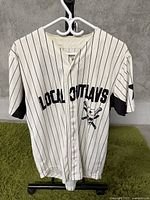Full front view of white baseball jersey with black pinstripes and Local Outlaws text and skull graphic.