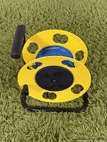 Large cord storage reel with blue cable and four black power outlets in the center, yellow circular sides with holes, black handle, placed on green carpet.