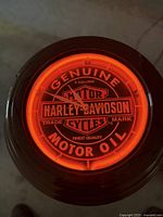 Photo of a round electric wall clock lit with bright orange neon. Features Harley-Davidson Genuine Motor Oil vintage branding on the clock face with white clock hands, black outer frame. Clock appears to be functioning light to illuminate but may need work.