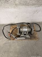 Full view of an electric bench grinder with two abrasive wheels, power cord, and base plate on a concrete floor.