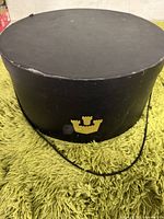 Black cylindrical vintage hat box with gold crown emblem. Shows scuffs and has a black string handle.