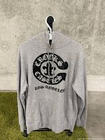 Front view of gray Crooks & Castles hoodie hanging on white hanger against concrete wall, showing logo and text details.