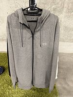 Front view of grey Oakley full-zip hoodie on hanger against concrete wall and green carpet.